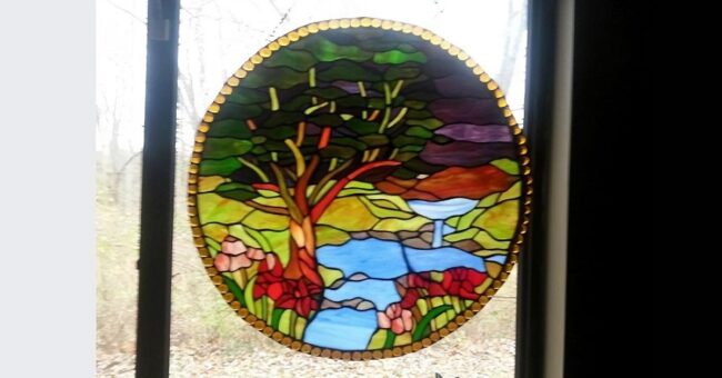 Photograph of a round stained glass depiction of a tree and brook, hanging in a window.