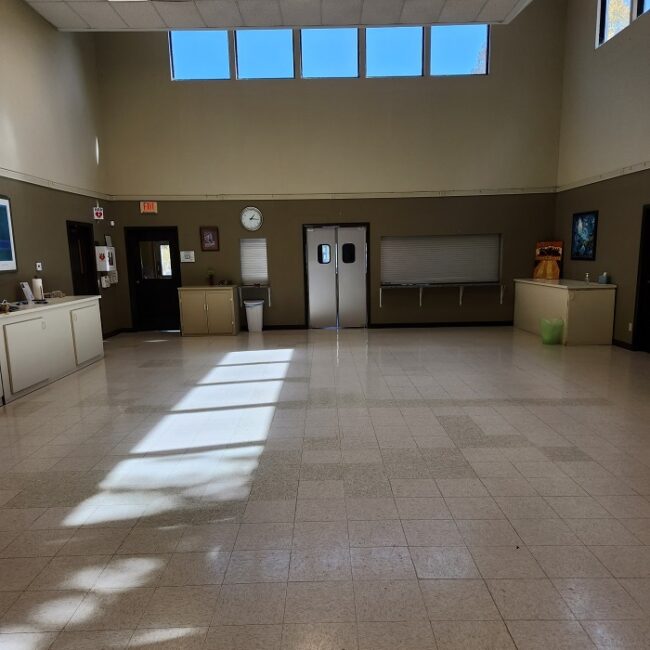 Large room with high ceiling. At the end are double-doors and a kitchen pass-through. The room has counters at the side but is relatively empty.