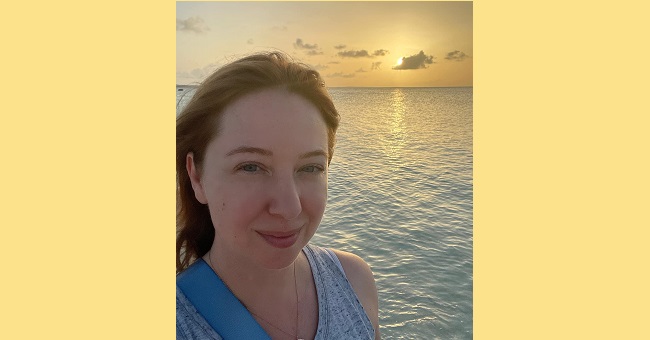 Woman looking at the camera while behind her is a sunset over water.