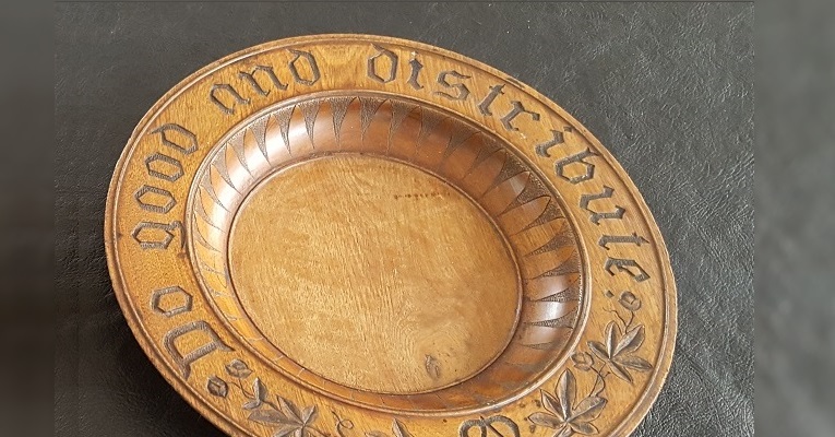 A wooden offering plate from over a century ago, carved with decoration and the sentence "Do good and distribute."