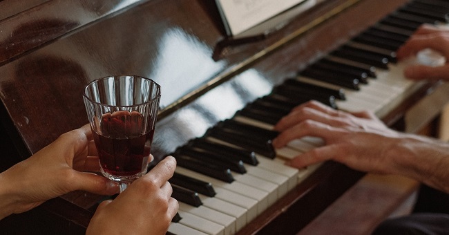 A person holds a wine glass while another plays the piano.