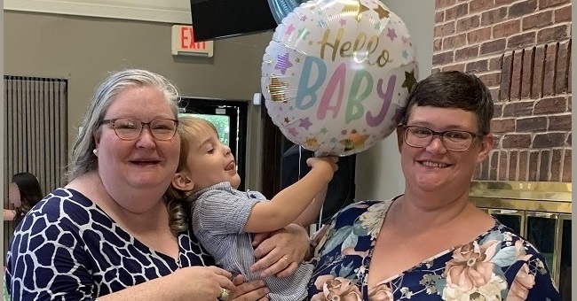 Two women hold a toddler, who holds several balloons, one of which has the word "Baby."
