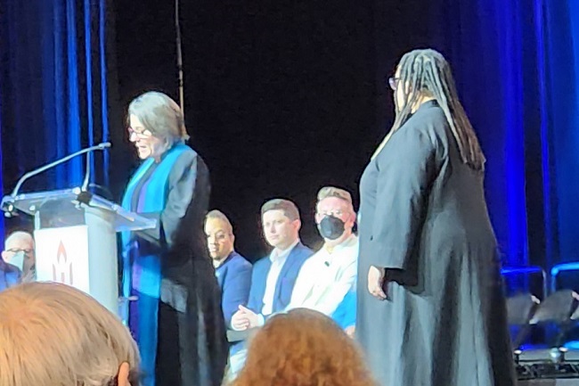 A woman in a blue robe stands reading at the lecturn, while another woman in a dark robe stands a couple paces back and to the side.