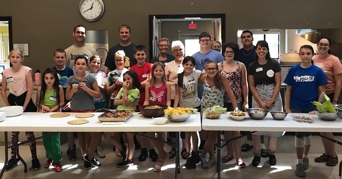 A large group of teens and pre-teens stand behind a table with food they have prepared.