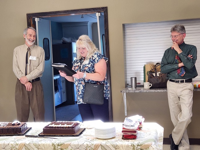 Three people stand behind a table with several cakes. The woman is reading to one of the men.