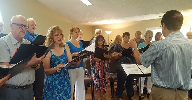 A choir of men and women stands as they rehearse, with the choir director leading them.